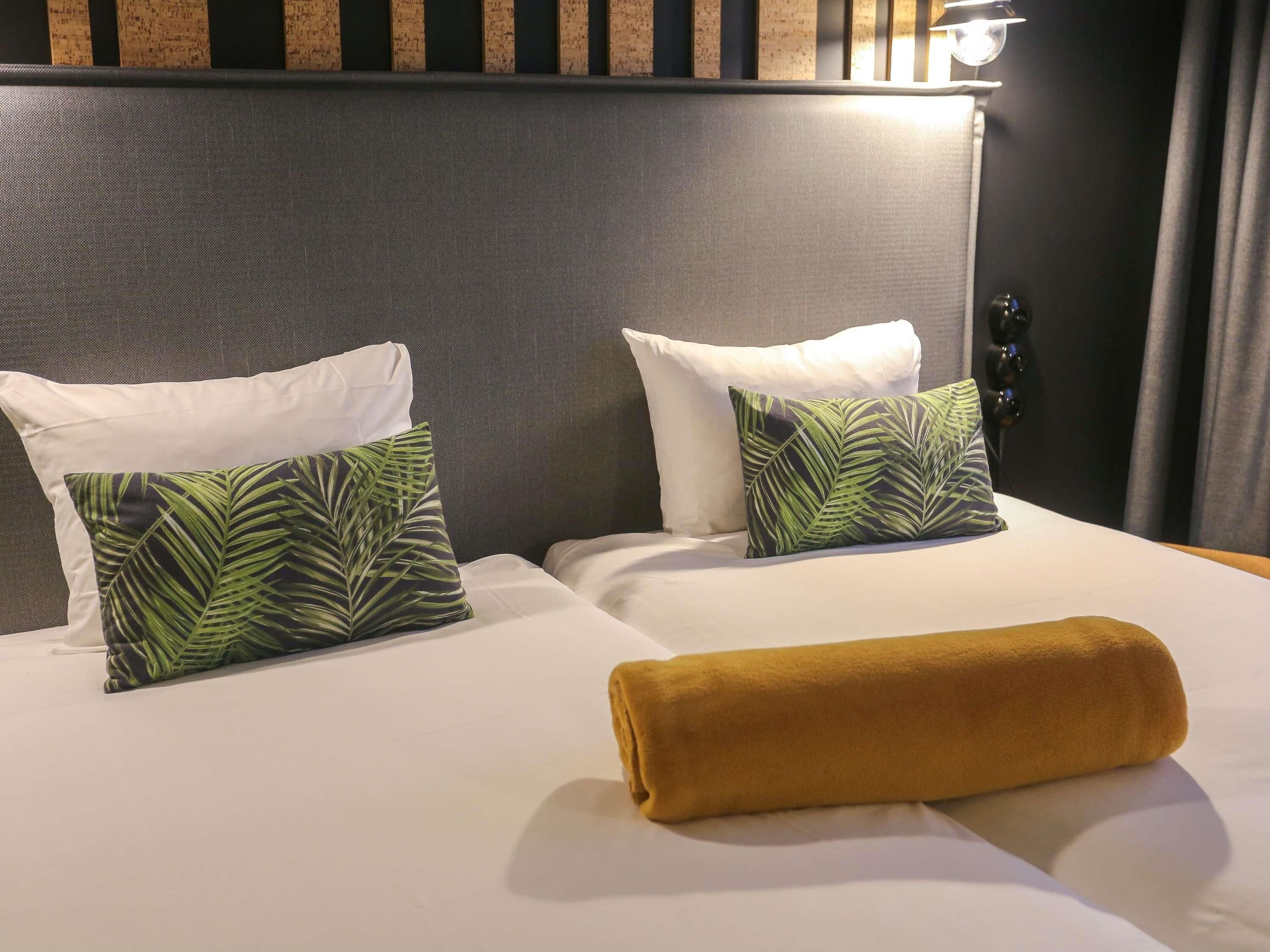 Ein Hotelzimmer mit einem Bett, zwei Kopfkissen mit Blättermotiv, einem weißen Kopfkissen und einem braunen Handtuch, sowie einem Kopfteil mit vertikalen Leisten.
