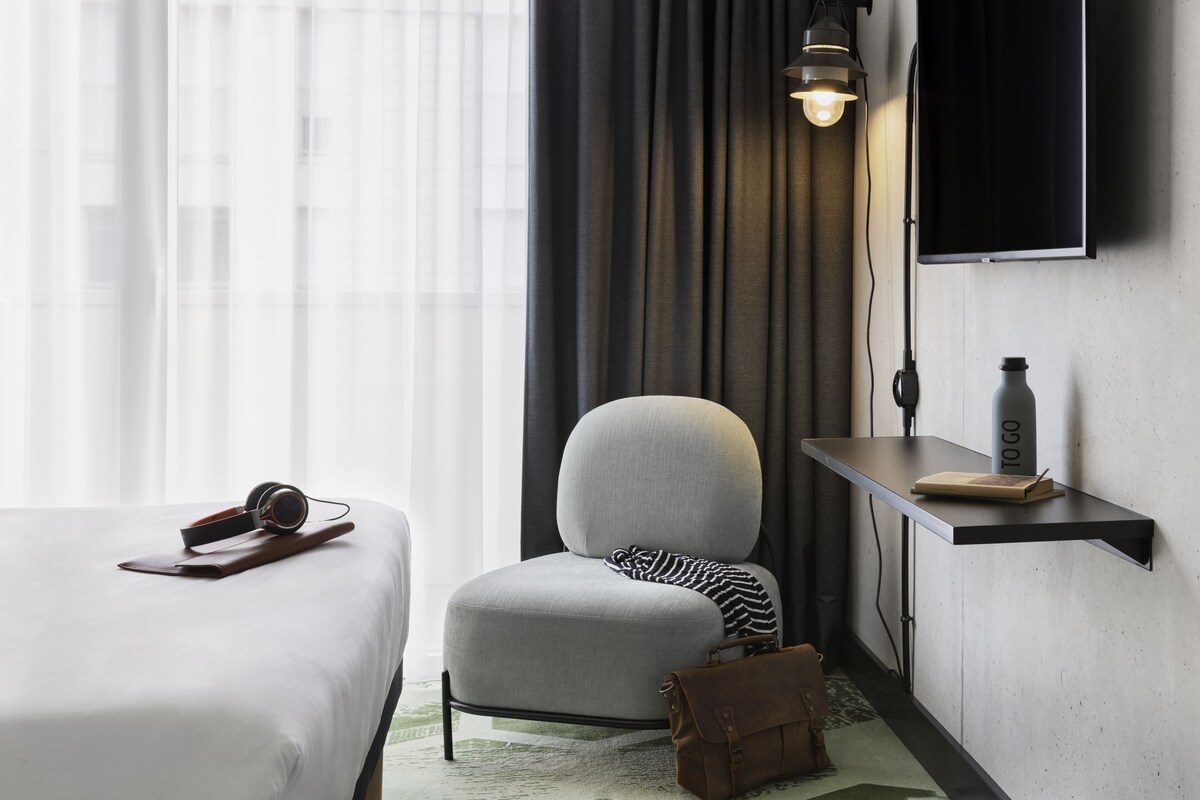 A hotel room with a bed, a chair, a desk, and a TV mounted on the wall.