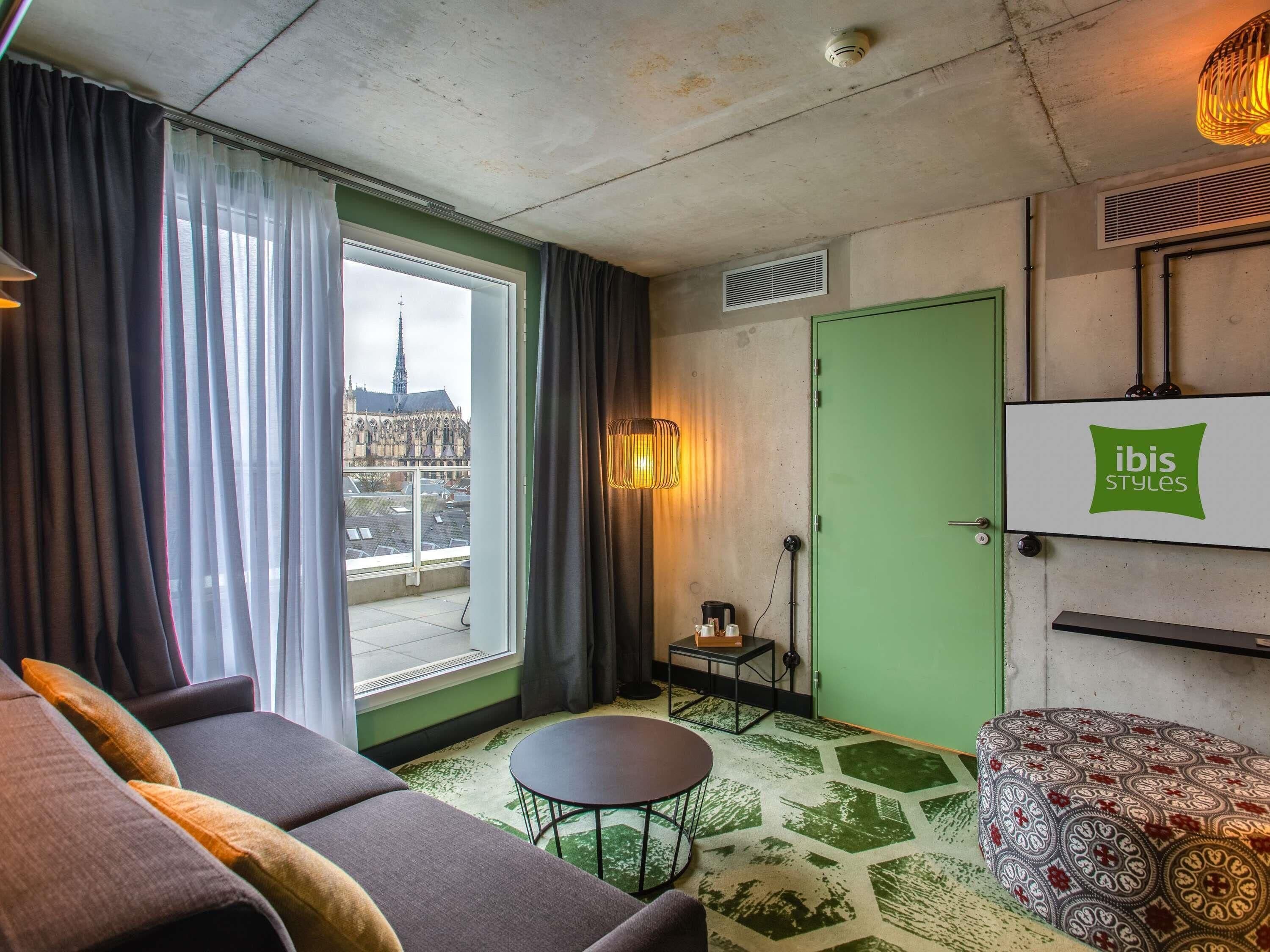 Une chambre d’hôtel avec un canapé, une petite table, un tapis à motifs, une porte verte et une vue sur une cathédrale par la fenêtre.