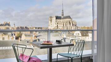 A balcony with a table, two chairs, a pair of glasses, and a camera. A pink jacket is draped over one chair. In the background, a large cathedral with a spire is visible.