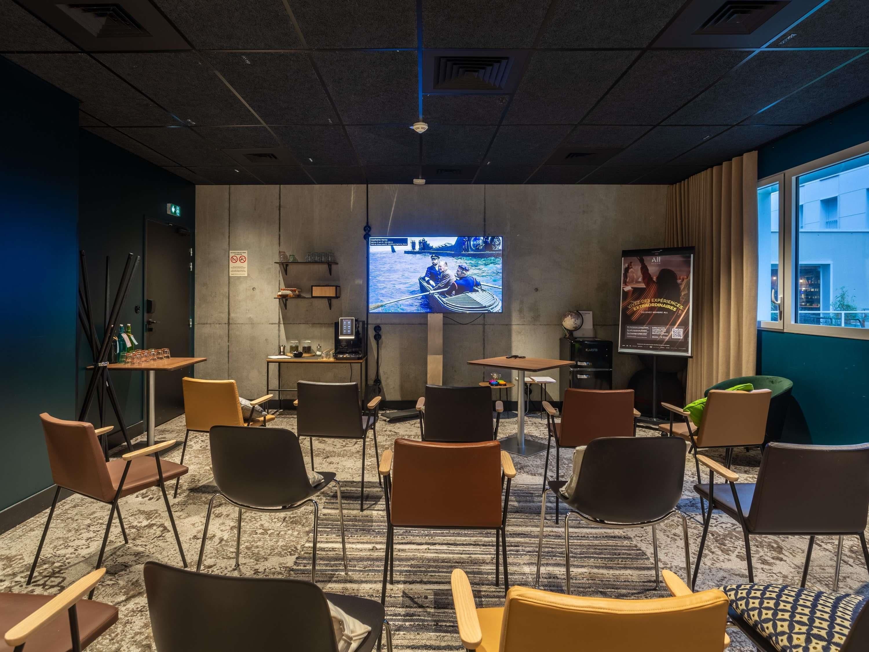 A conference room with a large screen, multiple chairs, tables, and a coffee machine.