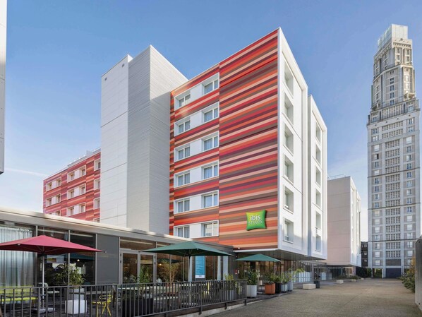 Un hotel moderno con fachada roja y blanca, un letrero verde de 'ibis styles' y un patio con mesas al aire libre.