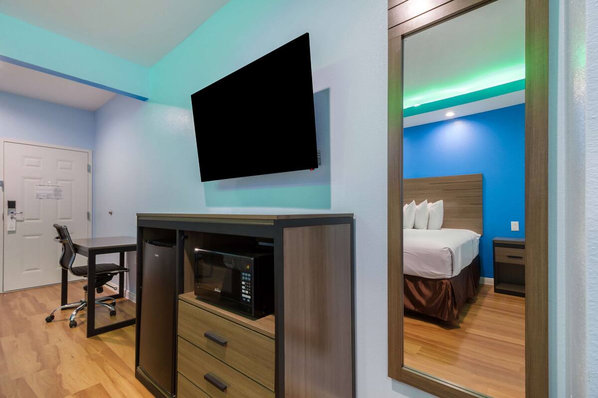 A hotel room with a flat-screen TV mounted on the wall, a wooden cabinet with drawers, a microwave, a desk with a chair, and a bed with a headboard.