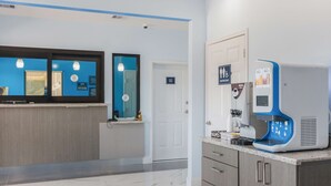 Zona de recepción de un hotel con una máquina de café, un cartel para los baños de empleados y un mostrador de recepción con un ordenador.