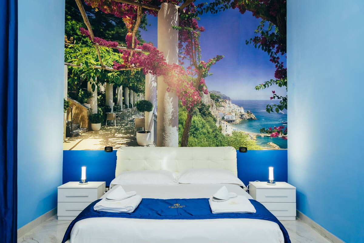 Une chambre à coucher comprenant un lit, deux tables de chevet et une grande fenêtre donnant sur une ville côtière et des falaises.