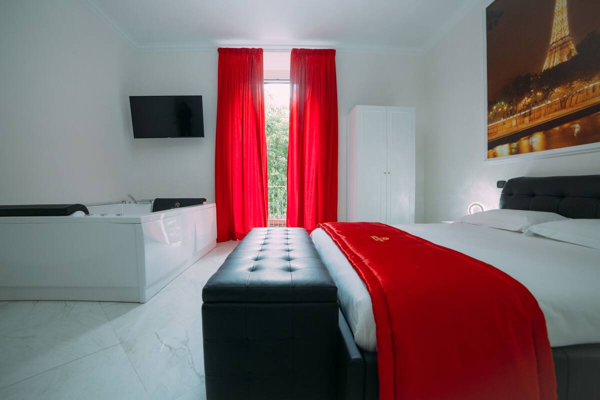 Une chambre d’hôtel moderne dotée d’un grand lit, d’un coin salon avec un canapé et une petite table, d’un téléviseur à écran plat fixé au mur et d’une fenêtre avec des rideaux rouges.