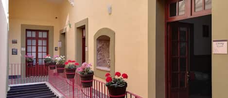 A building with multiple doors, some labeled as "Living Room" and "Café." There are potted plants with flowers along the railing of a staircase.