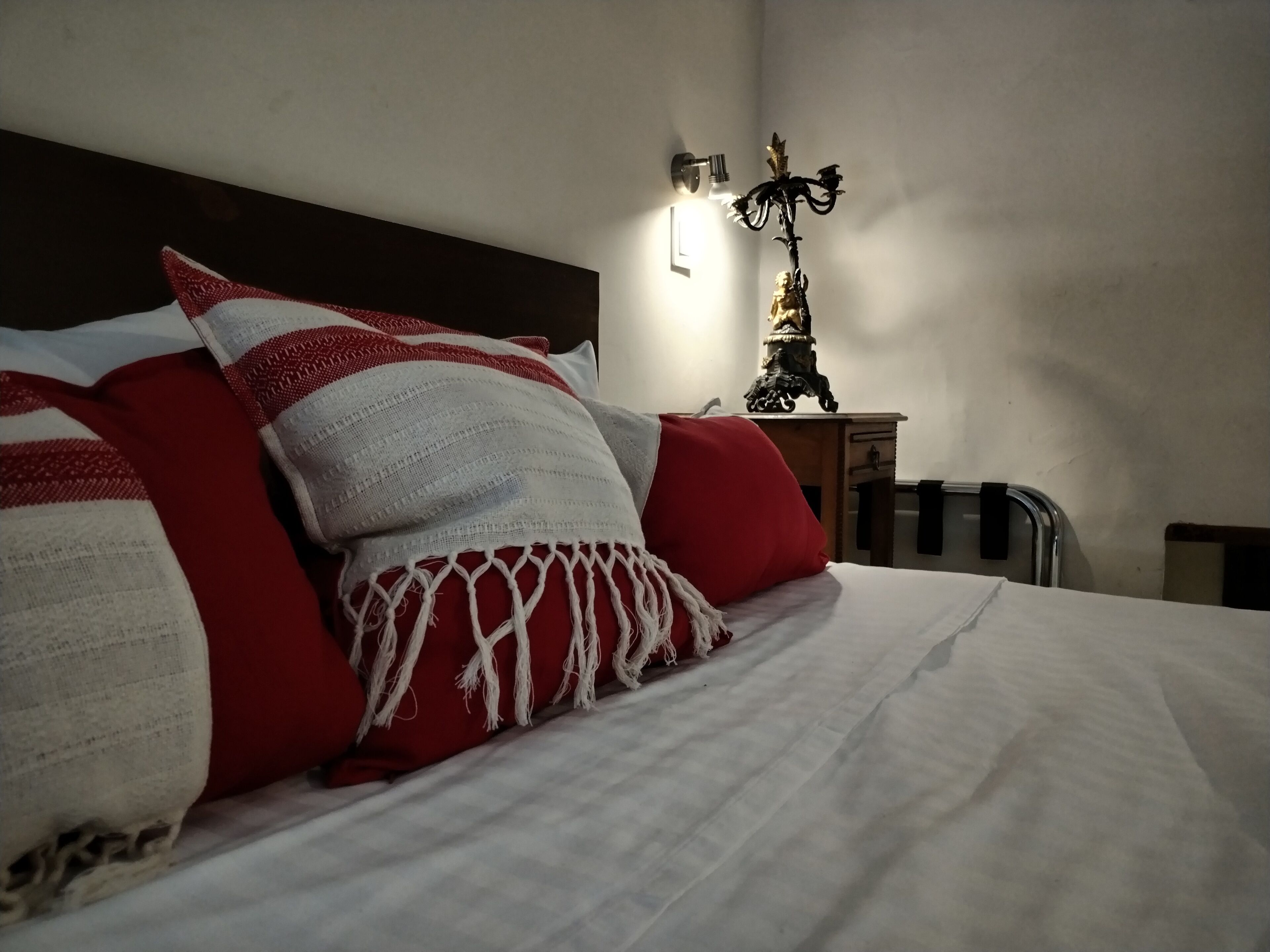 Una cama cuidadosamente tendida con ropa de cama roja y blanca, una lámpara de pared y una pequeña mesa con un objeto decorativo.