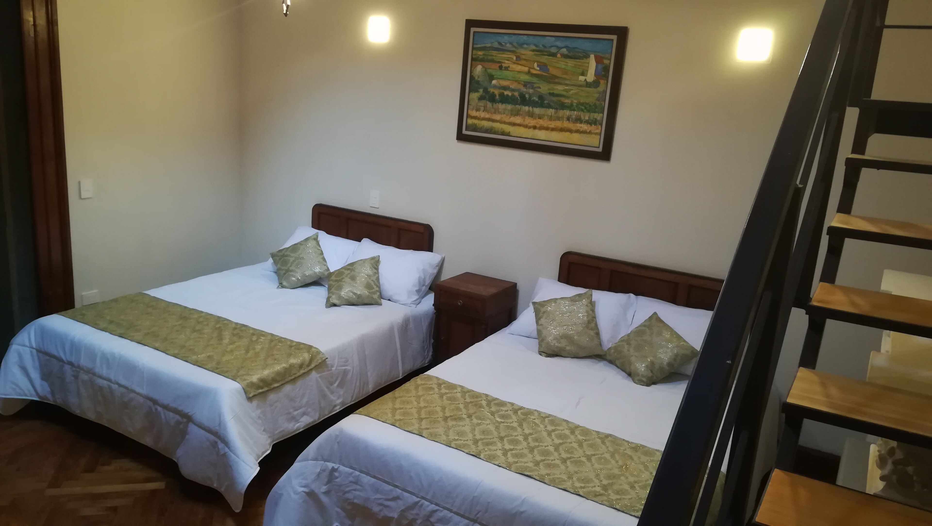 Habitación con dos camas, cada una con cabecero de madera, ropa de cama blanca y almohadas decorativas. Hay un cuadro en la pared sobre las camas y una escalera de madera a la derecha.