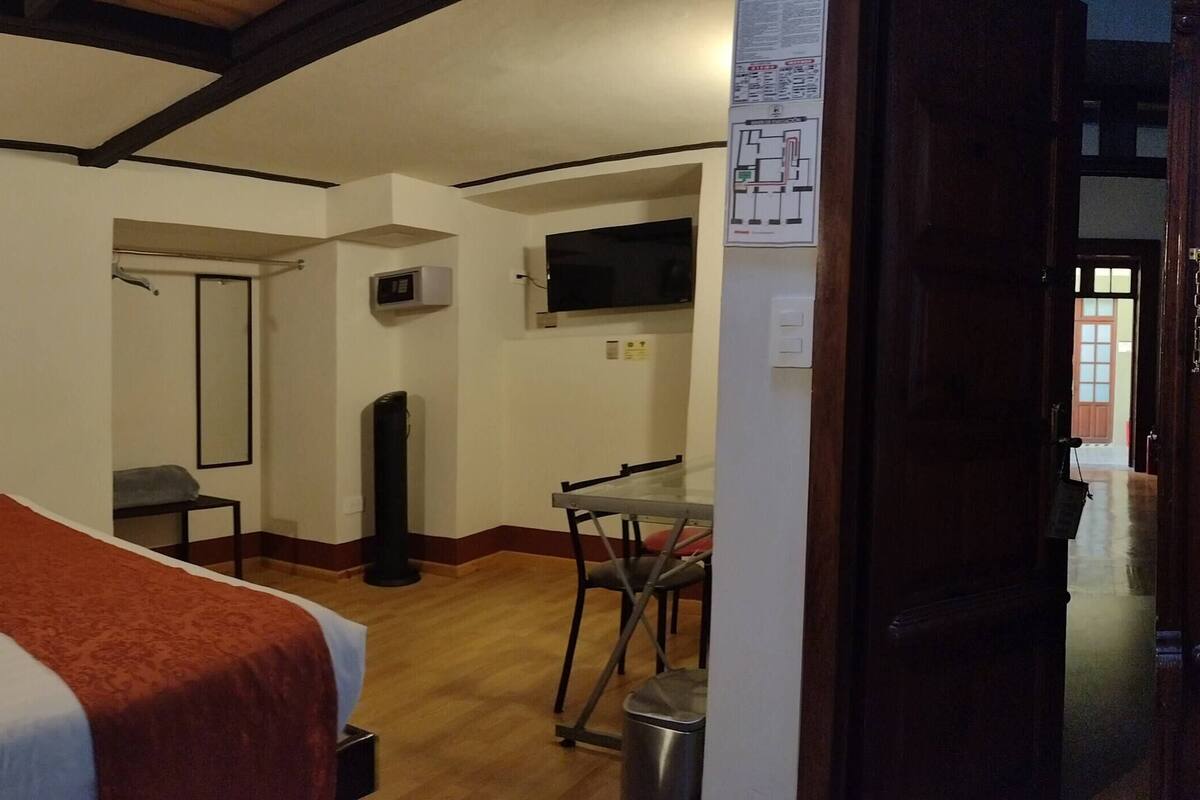 Quarto de hotel com cama, escrivaninha, cadeira, televisão e porta que dá para outro cômodo.