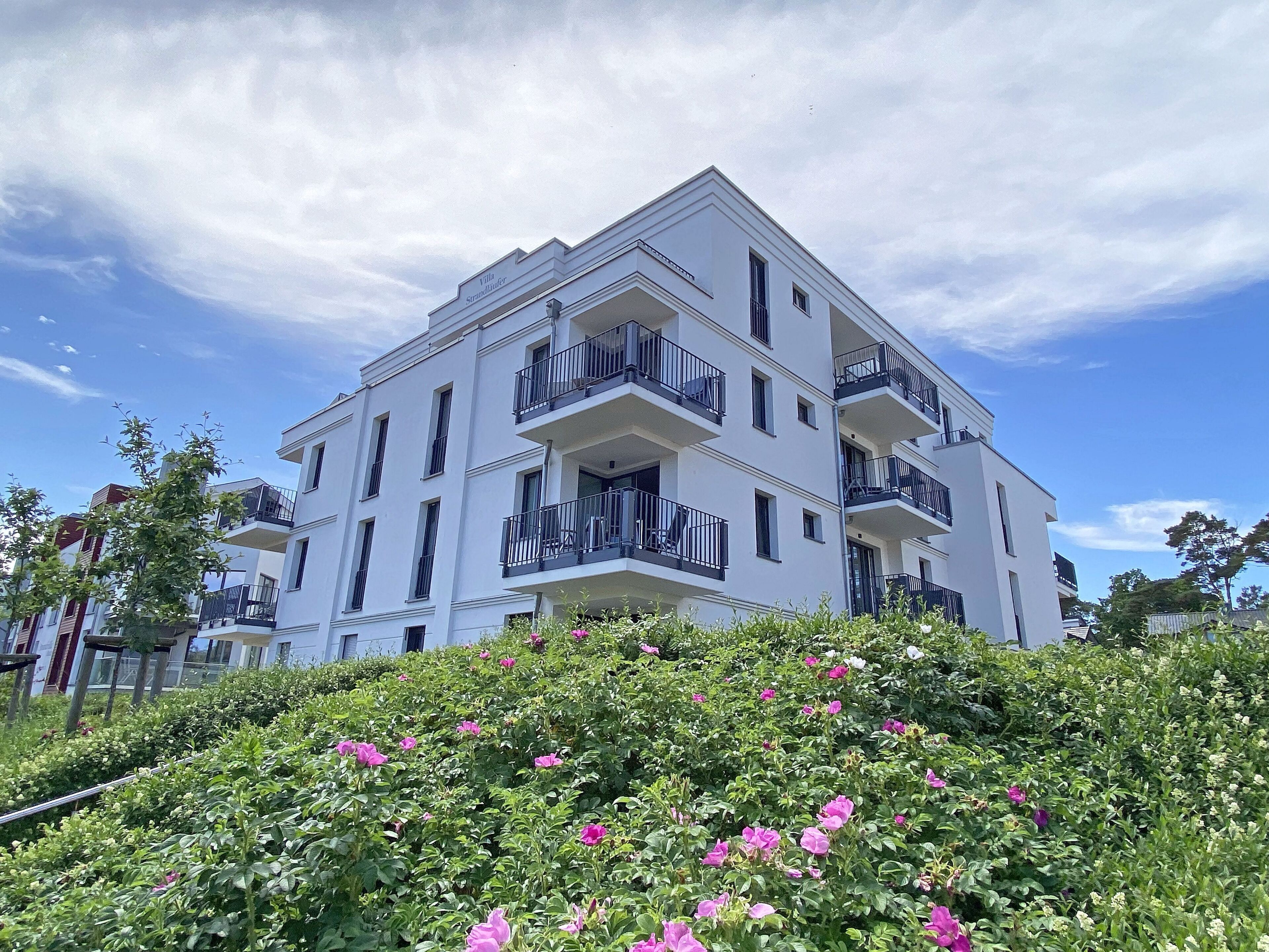 Un immeuble blanc à plusieurs étages, doté de balcons, entouré d’une végétation luxuriante et de fleurs roses.