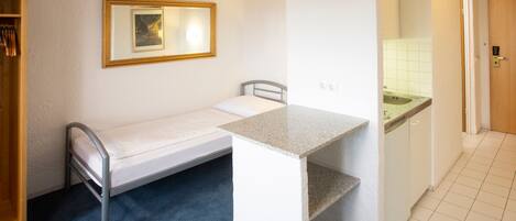 A compact room with a single bed, a small table, a mirror, and a kitchenette area.