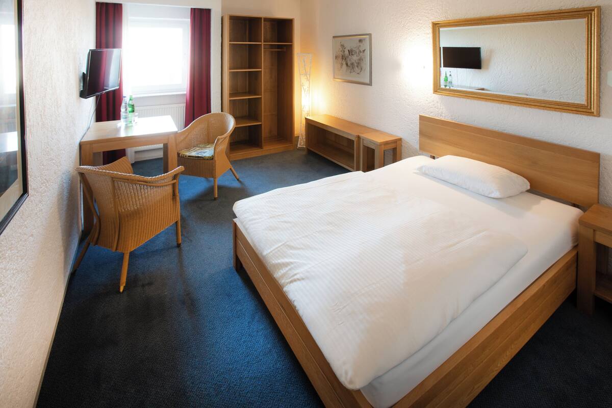 Ein Hotelzimmer mit Bett, Schreibtisch mit Stuhl, Kleiderschrank, Nachttisch und einem an der Wand befestigten Fernseher.