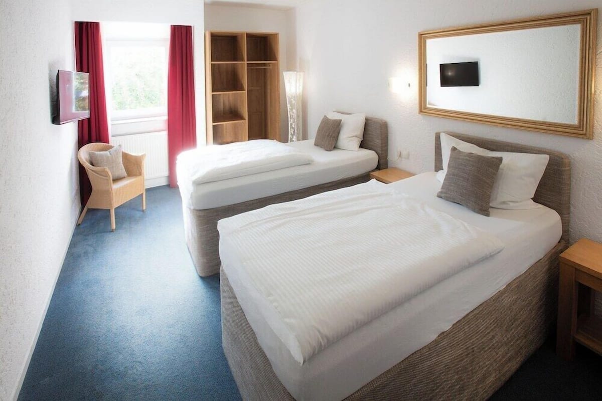 Ein Hotelzimmer mit zwei Betten, einem Kleiderschrank, einem Sessel und einem an der Wand befestigten Fernseher.