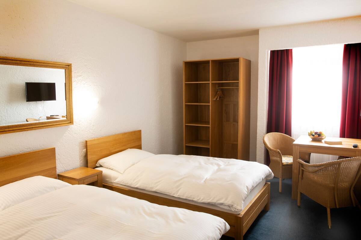 Ein Hotelzimmer mit zwei Betten, einem Fernseher, einem Kleiderschrank, einem Schreibtisch mit Stuhl und einem fenster mit Vorhängen.