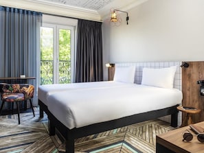 A hotel room with a bed, a chair, a small table, and a view of greenery outside.