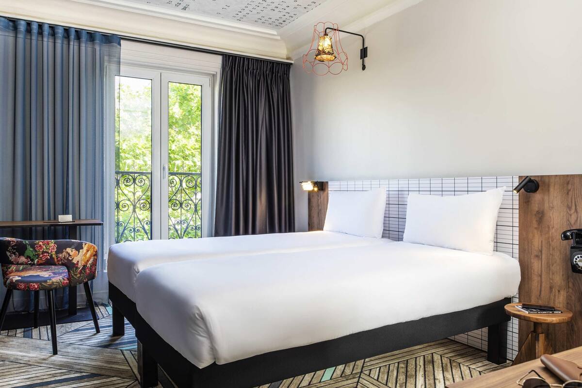 A hotel room with a large bed, a wooden nightstand, a chair with a floral pattern, and a view of greenery through the window.
