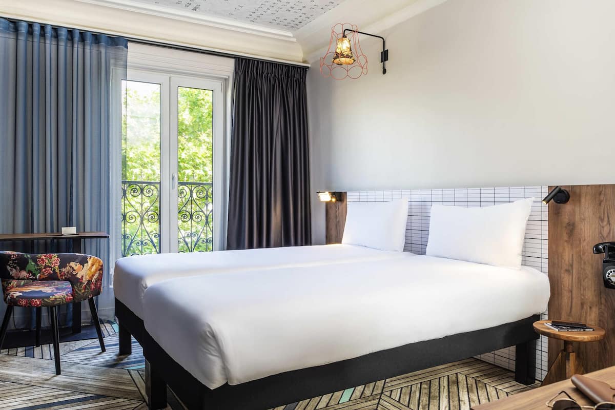 Une chambre dâhĂŽtel avec un grand lit, une table de chevet en bois, une chaise Ă motifs floraux et une vue sur la verdure par la fenĂȘtre.