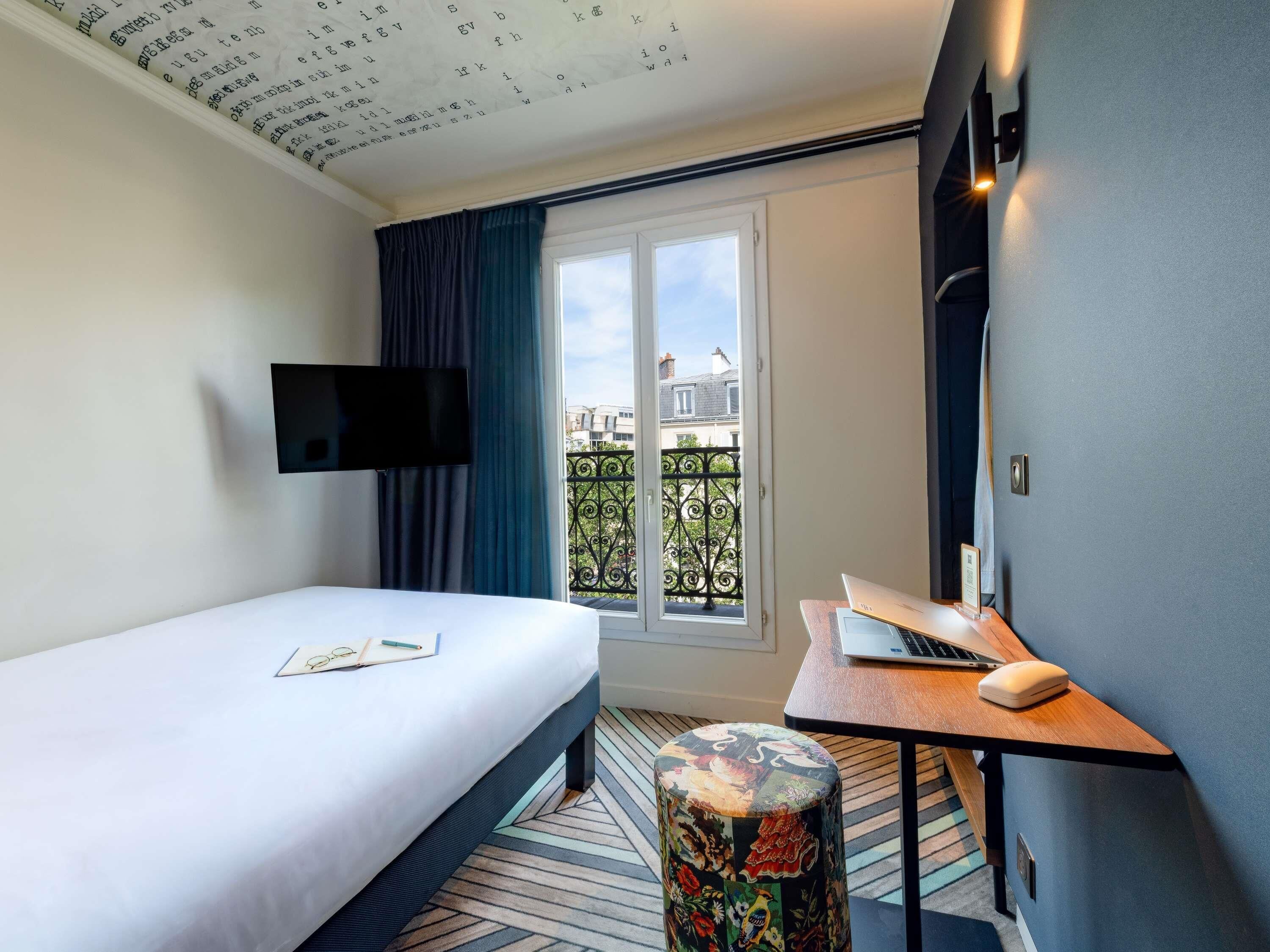 A hotel room with a bed, a desk with a laptop, a small stool, a TV mounted on the wall, and a window with a balcony view.