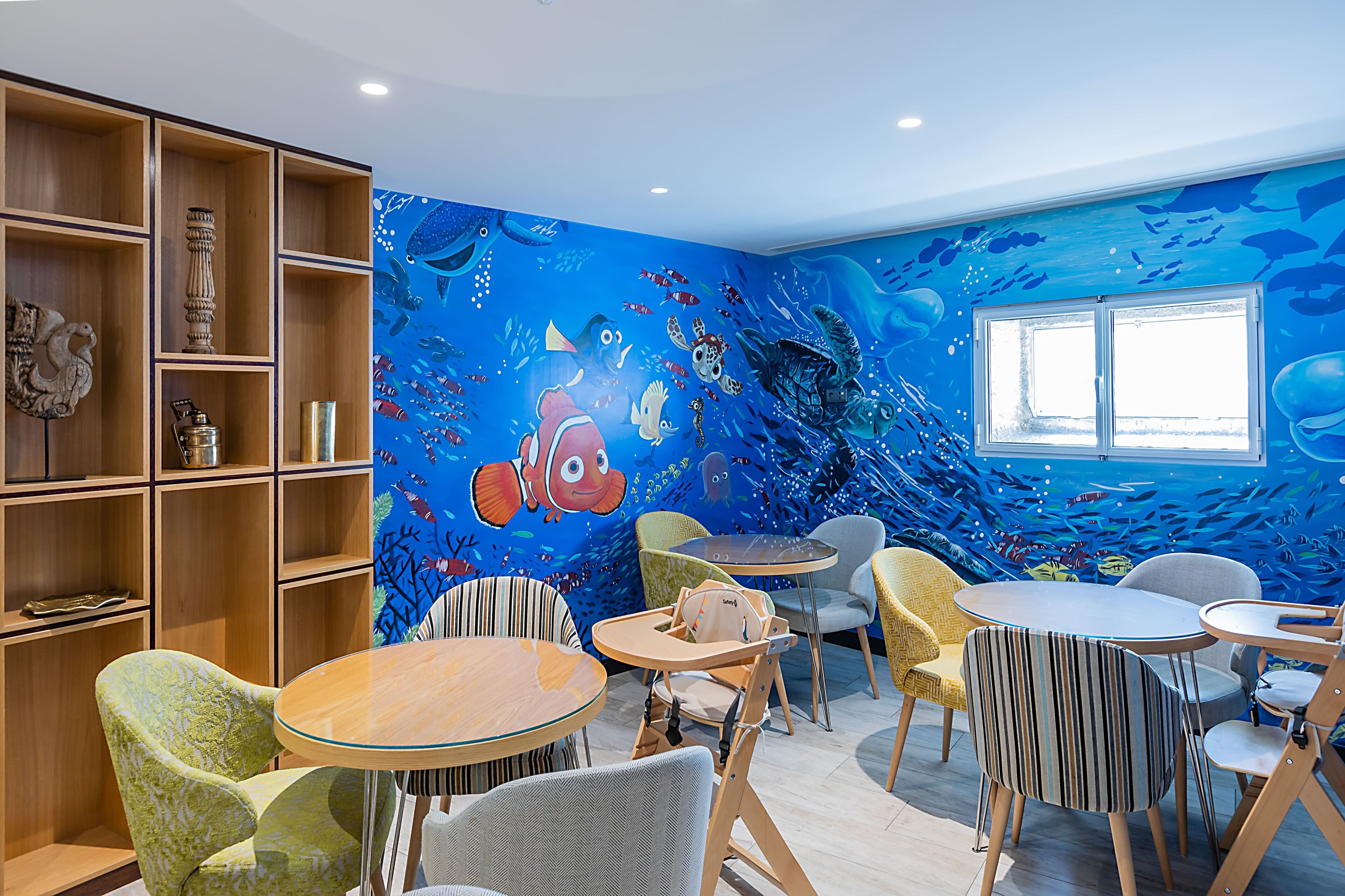 A children's playroom with a mural of underwater scenes, wooden shelves, and various seating arrangements.