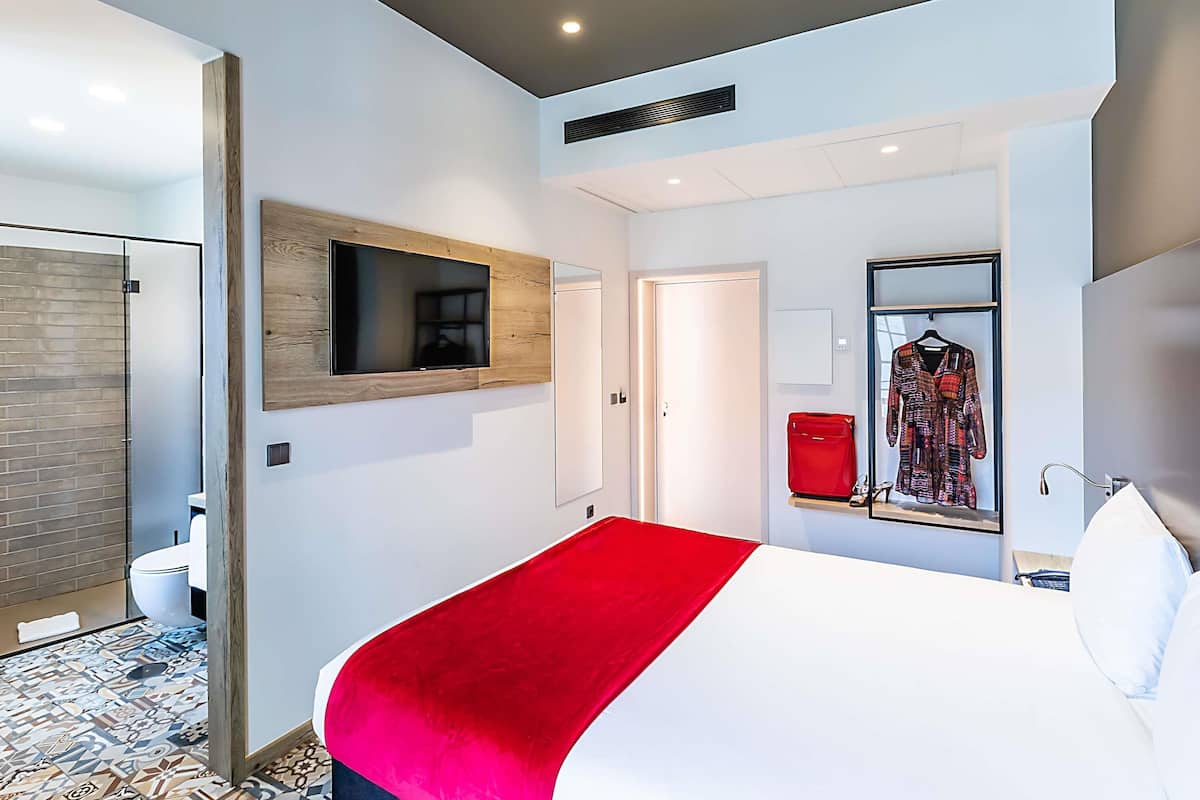 Una habitación de hotel moderna con una cama grande, un televisor montado, un armario y un baño visible a través de una puerta abierta.