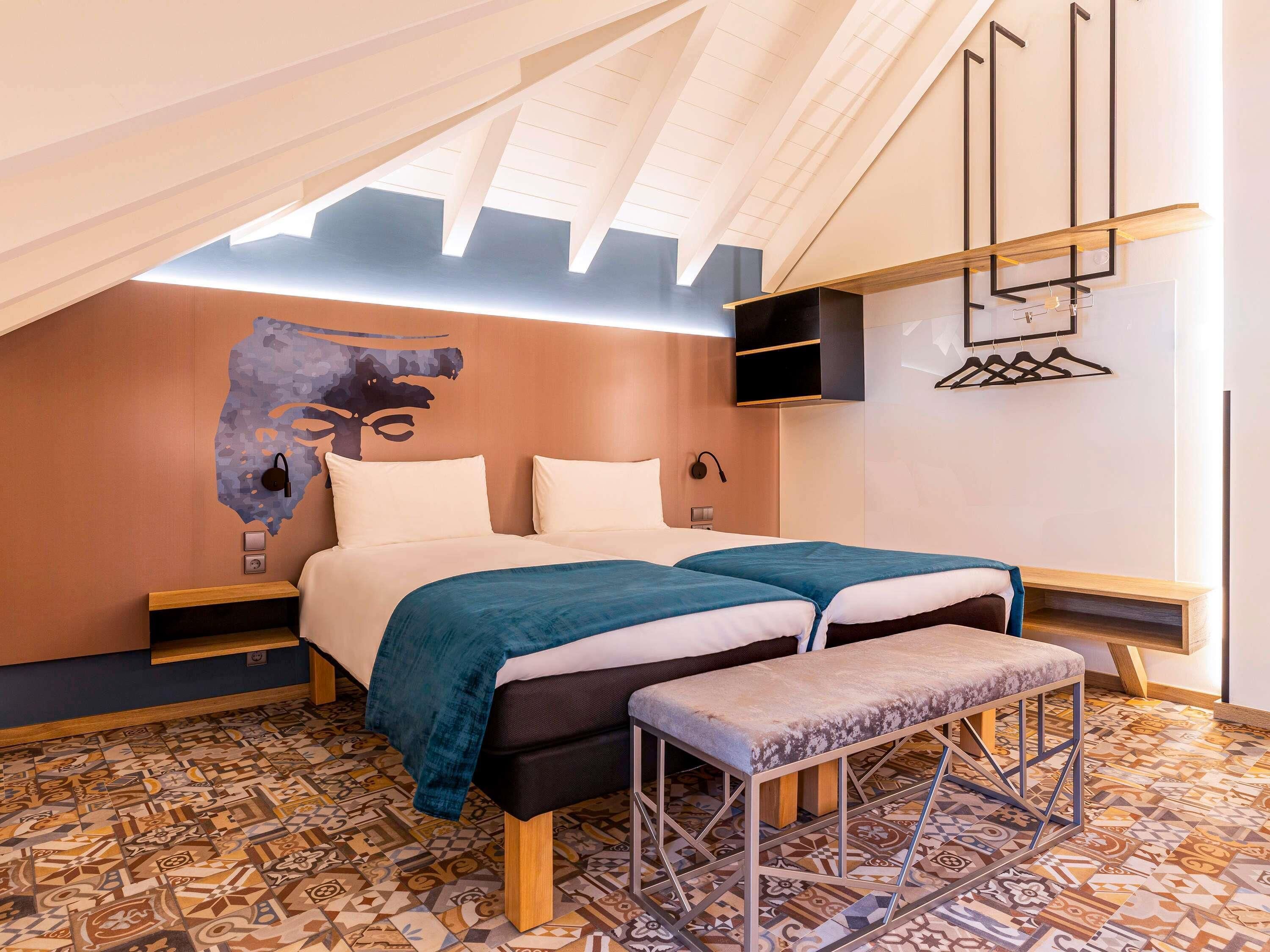 A modern bedroom with a sloped ceiling, a large bed with two pillows, a bench, and a wall-mounted coat rack.