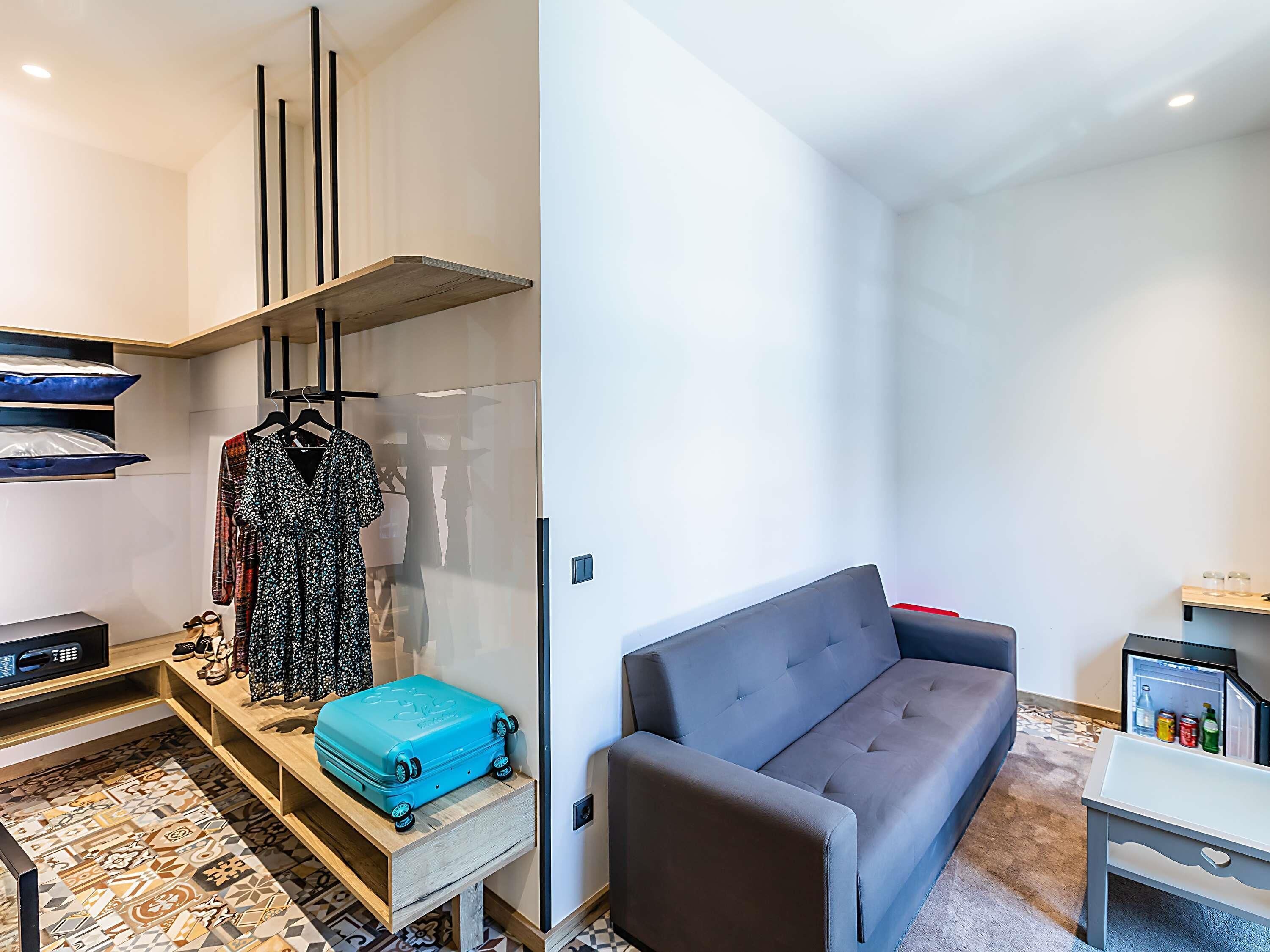 A compact living space with a sofa, a small table, a blue suitcase, and a wall-mounted shelf with a dress and sunglasses.