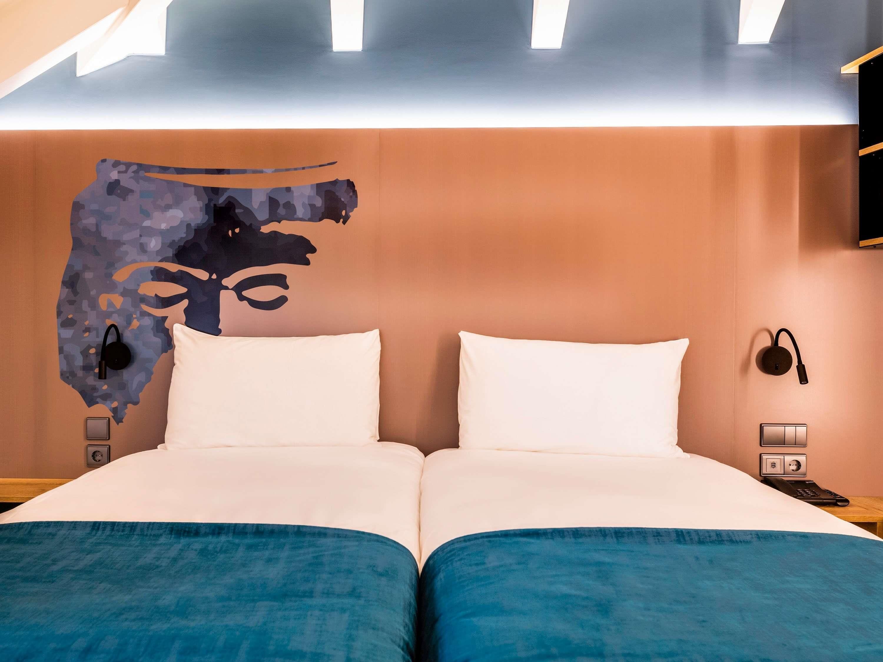 A double bed with white and blue bedding, a headboard with a camouflage pattern, a wall-mounted phone, and a light fixture above the bed.