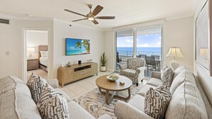 A spacious living room with a ceiling fan, a large window with a view of the ocean, a dining area with a table and chairs, and a cozy seating area with patterned cushions. - Beachfront Condo With Huge Views/pools/tennis/grills (Marco Island)
