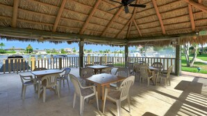 Un espace extérieur abrité, aménagé avec des tables et des chaises, doté d’un ventilateur de plafond et offrant une vue sur l’eau et la végétation.