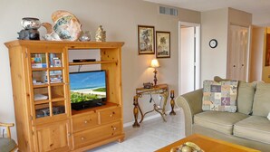 Un salon comprenant un meuble de rangement en bois pour l’équipement audiovisuel, un téléviseur, une table avec un panier de fruits, un canapé et une lampe.
