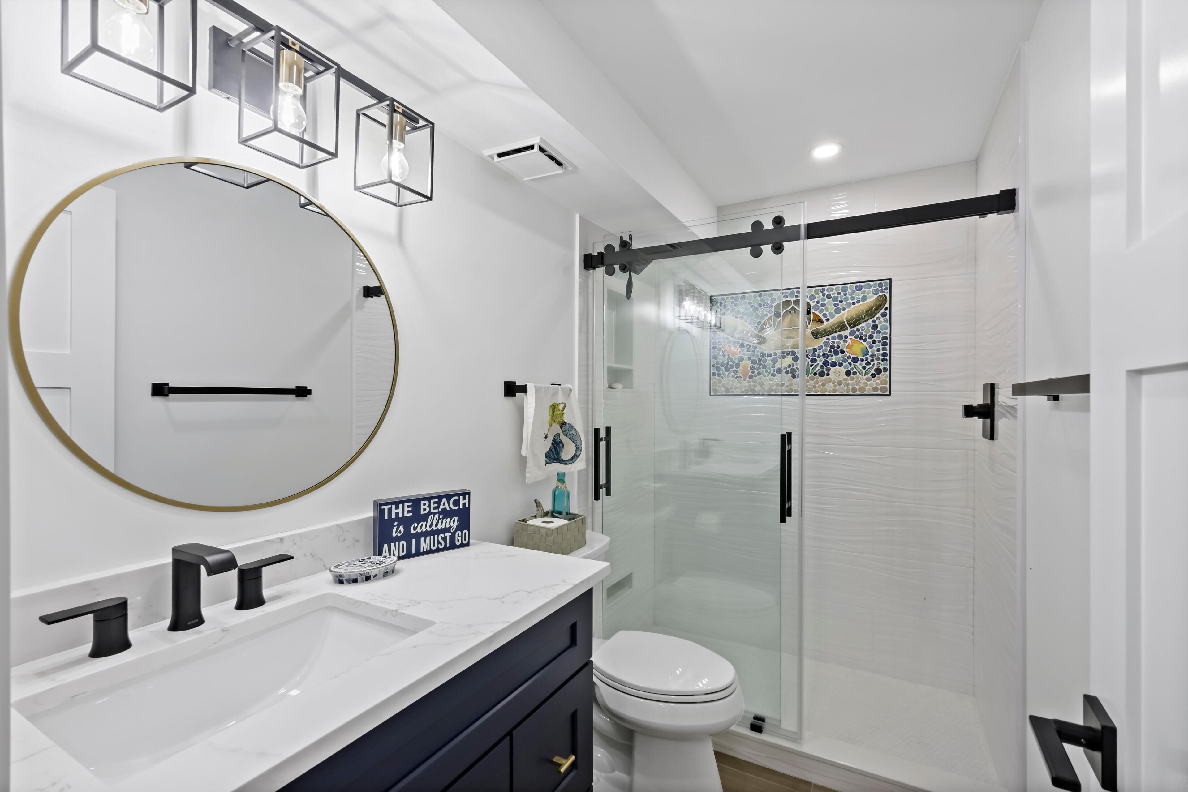 A modern bathroom with a white sink, black faucets, a large round mirror, and a glass-enclosed shower.