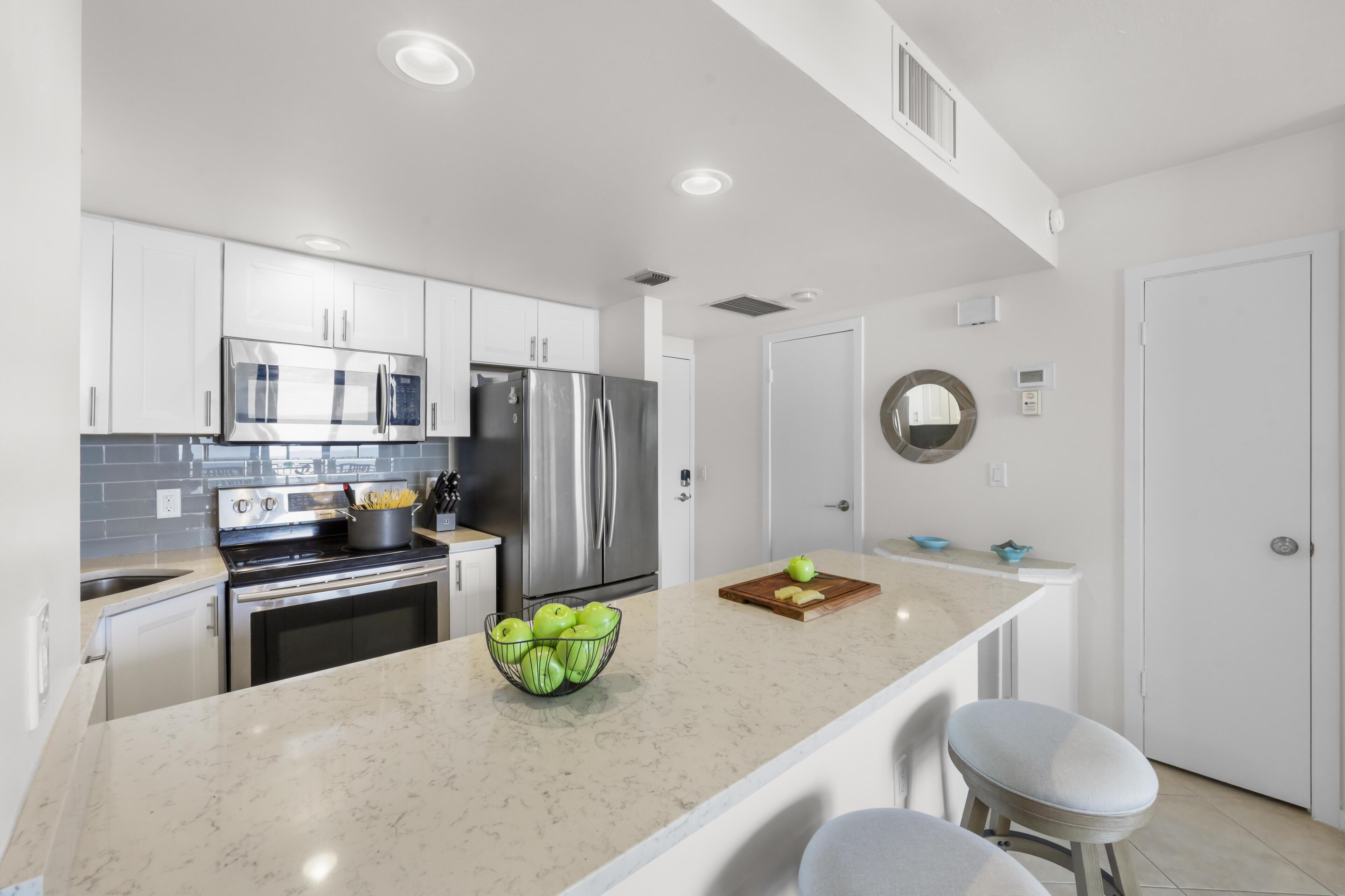 A modern kitchen with white cabinets, stainless steel appliances, a marble countertop, and a bowl of green apples.