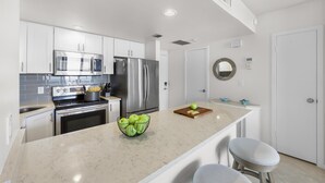 A modern kitchen with white cabinets, stainless steel appliances, a marble countertop, and a bowl of green apples. - Tradewinds 701 (Marco Island)