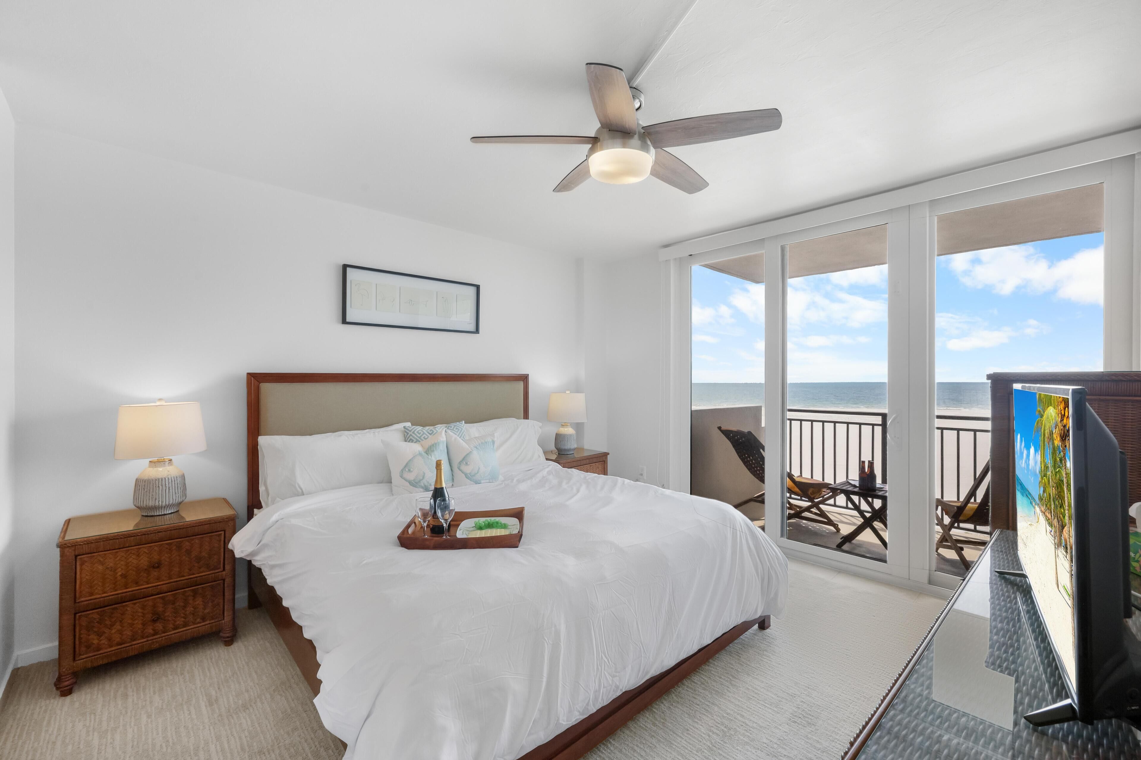 A bedroom with a large bed, a ceiling fan, a nightstand with a lamp, a tray with a bottle, and a painting on the wall.