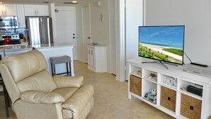 A modern living room with a leather armchair, a flat-screen TV on a white entertainment unit, and a kitchen in the background. - Tradewinds 701 (Marco Island)