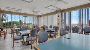 A spacious indoor dining area with multiple tables and chairs, large windows with blinds, ceiling fans, and a view of the outdoors. - Tradewinds 701 (Marco Island)