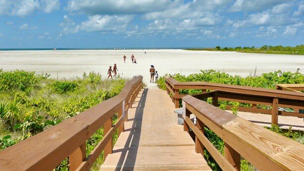 Une promenade en bois mène à une plage où l’on voit des gens se promener, et le ciel est dégagé.
