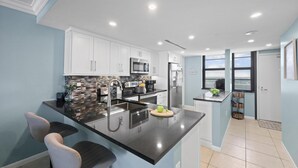 A modern kitchen with white cabinets, stainless steel appliances, a black countertop, and a large island with bar stools. - Beachfront Condo With Pool and Balcony Facing Beach (Marco Island)