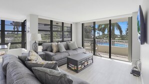 Ein modernes Wohnzimmer mit einem grauen Sofa, einem Couchtisch mit Büchern und großen Fenstern mit Blick auf einen Pool und Palmen.