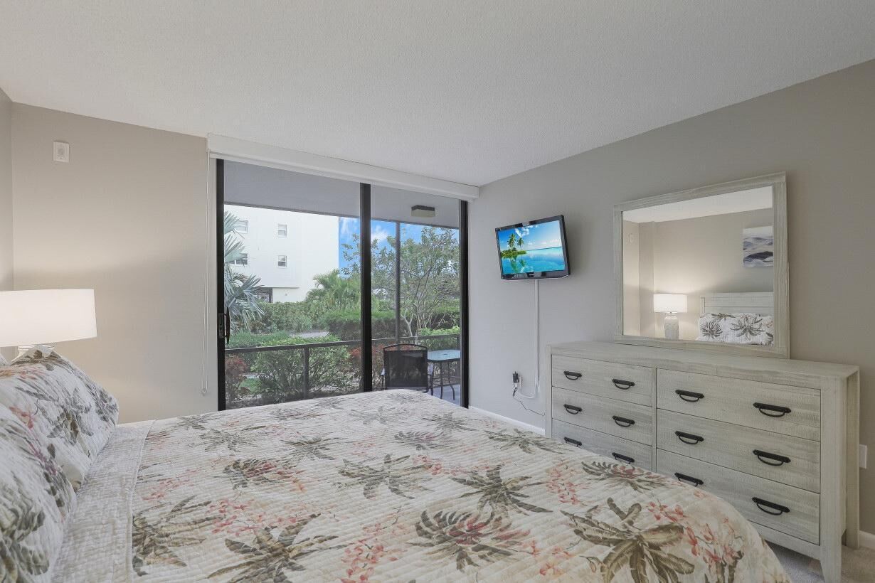 A bedroom with a bed, a dresser, a TV, and a balcony with a view of greenery and buildings.