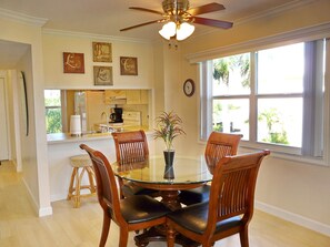 A dining area with a glass table surrounded by wooden chairs, a ceiling fan, and a window with a view of greenery. - Bright Condo in Community With Pool/steps to Beach (Marco Island)