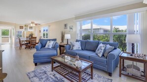 A spacious living room with a blue sofa set, a glass coffee table, and a dining area with wooden furniture. - Bright Condo in Community With Pool/steps to Beach (Marco Island)