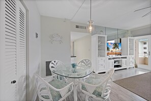 A dining area with a glass table and white chairs, a white cabinet with glass doors, and a large mirror reflecting a room with a TV and a beach scene.