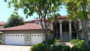 A two-story house with a red-tiled roof, a garage, and a well-maintained front yard with trees and shrubs. - Quiet, Gated Community With Pool and Tennis Courts (Marco Island)