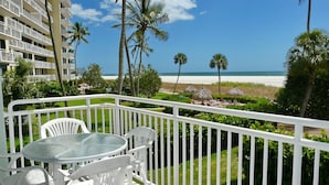 Un balcon avec une table en verre et des chaises blanches, donnant sur une plage bordée de palmiers et sur un bâtiment d'hôtel en arrière-plan.