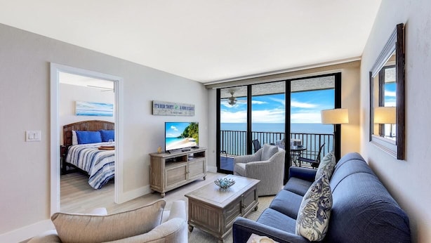 A modern living room with a sofa, armchairs, a coffee table, and a TV. There is a balcony with a view of the sea.