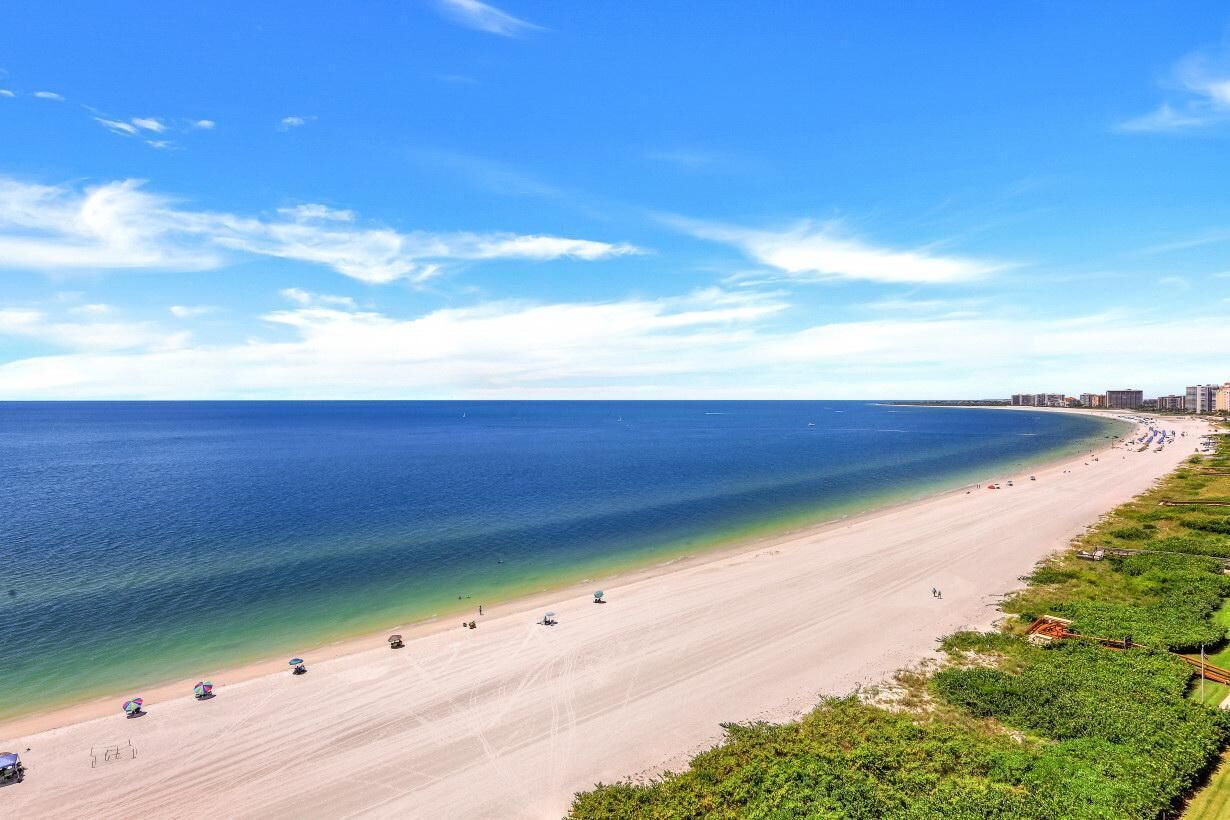 A wide beach with clear blue water, a sandy shore, and a line of buildings in the distance.