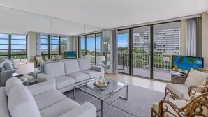 A spacious living room with a large sofa, a coffee table, and a television on a balcony overlooking a cityscape.