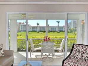 Een balkon met een glazen deur, witte stoelen, een tafel met een bloevas, en uitzicht op een verzorgd gazon en gebouwen op de achtergrond.